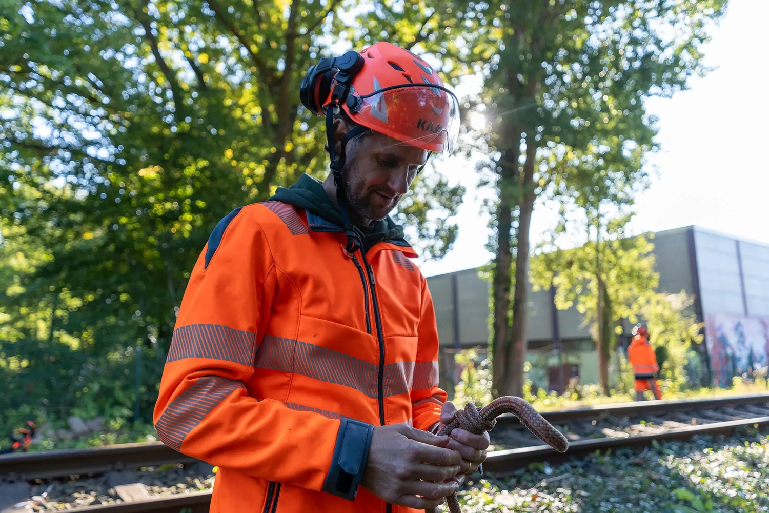 rundum baum GmbH - Baumpflege - Ein Arbeiter in orangefarbener Sicherheitsjacke und Helm hält einen Anschlagmittel-Schäkel in der Nähe von Bahngleisen.