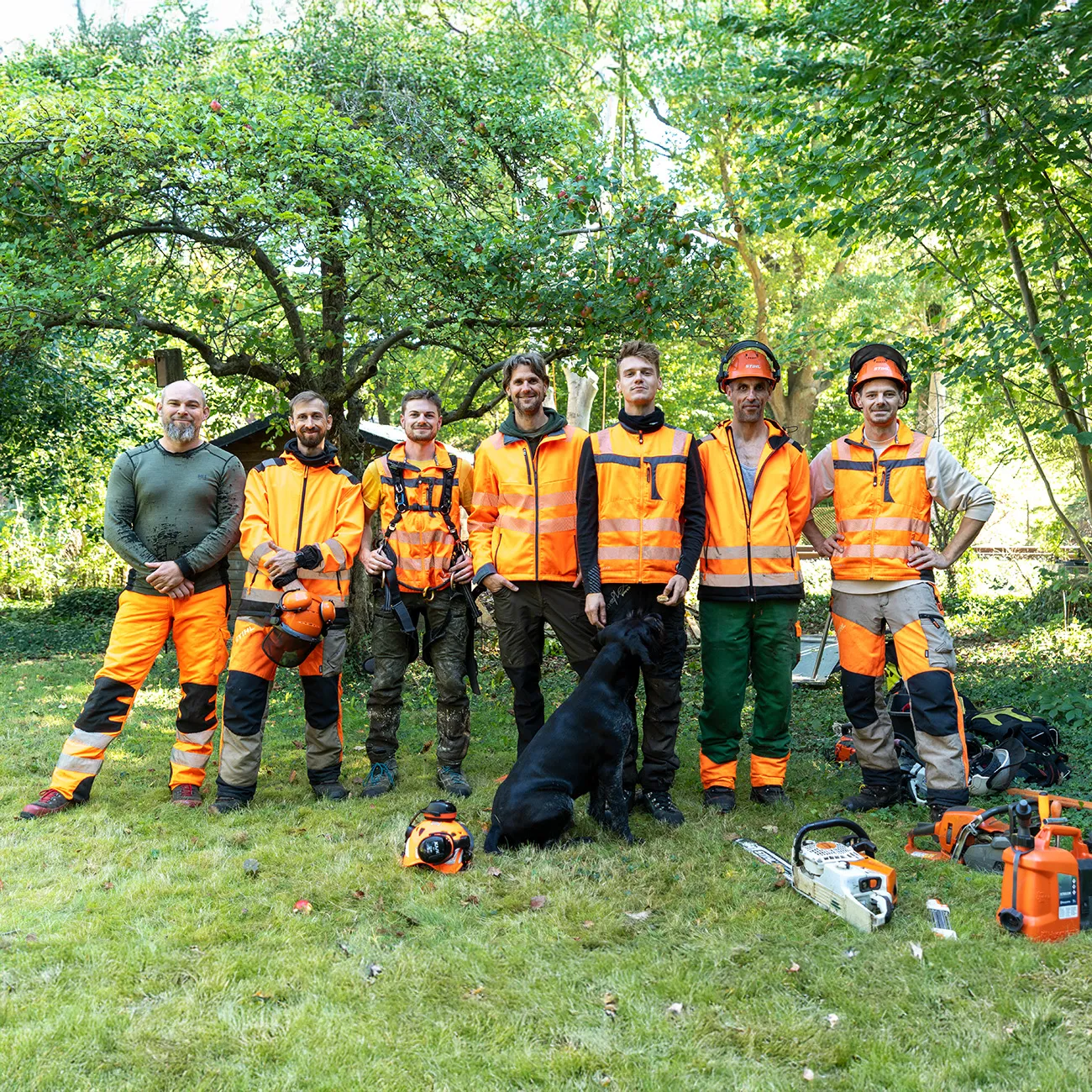 rundum baum GmbH - Baumpflege - Sieben Baumpfleger in Schutzkleidung posieren im Freien mit Kettensägen.